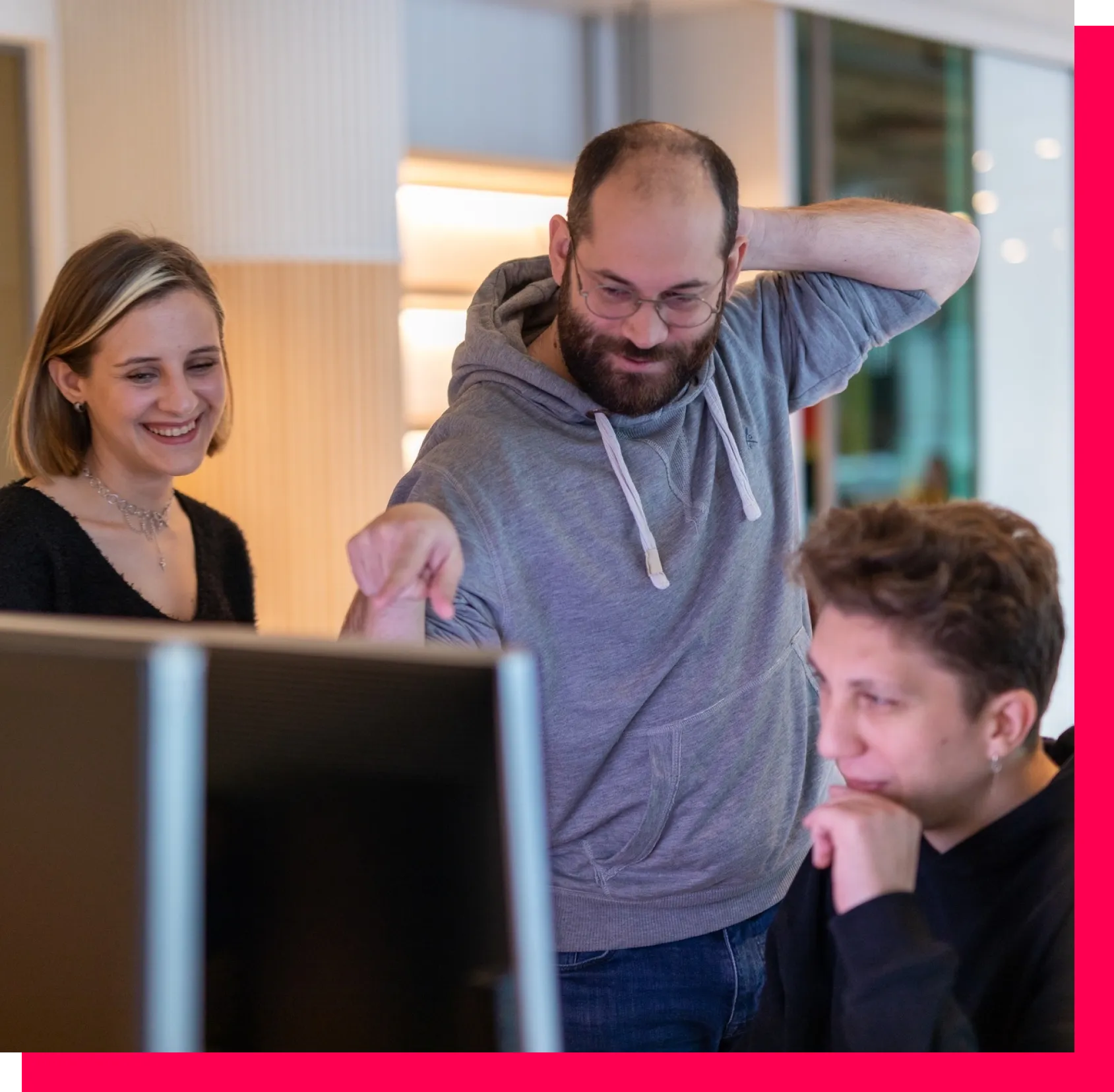 Three team members gathered in front of a large monitor, with one person pointing and the others looking engaged.
