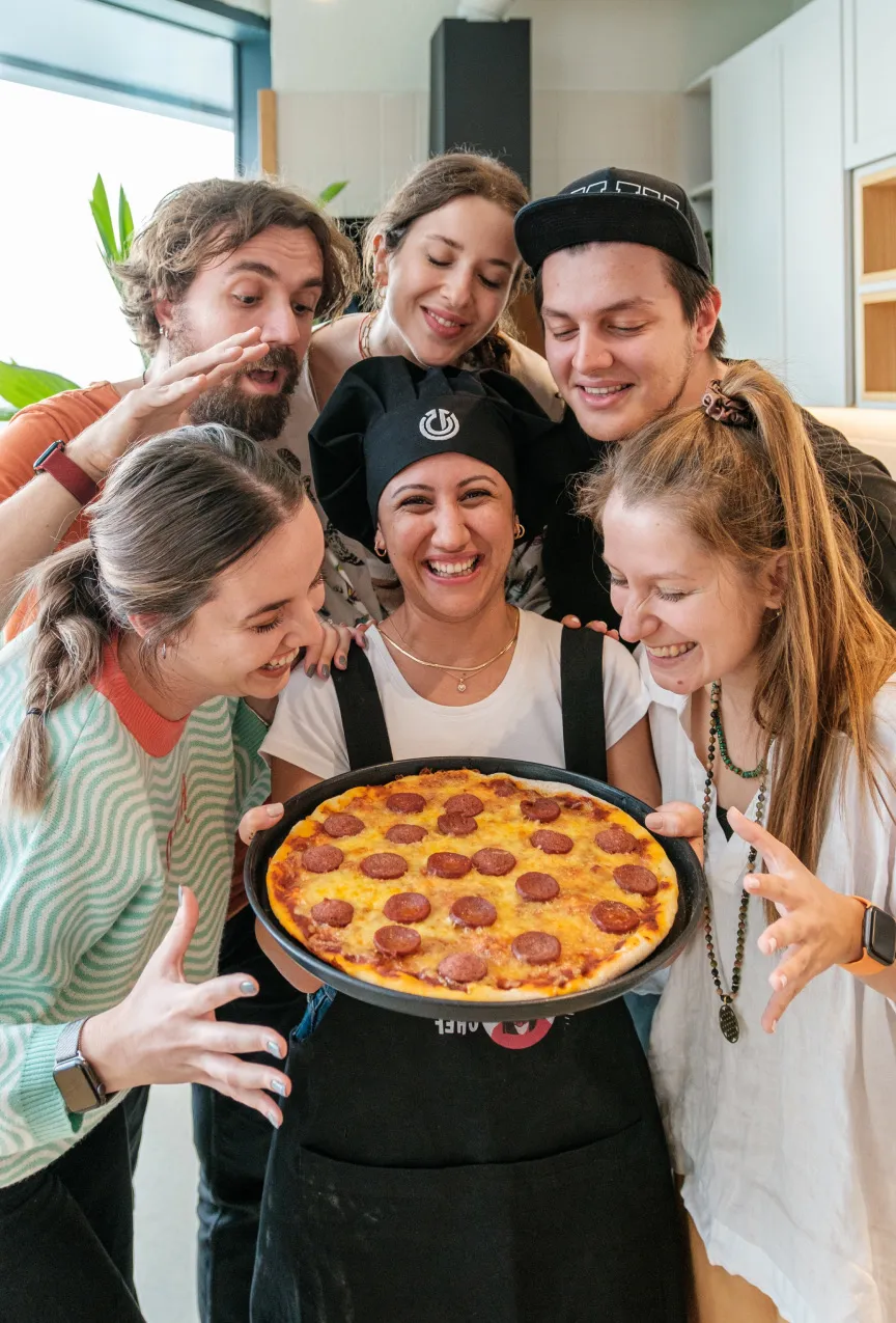 Chef Seço in a black kitchen apron and a black chef hat, holding pepperoni pizza surrounded by happy Atolye15 employees.