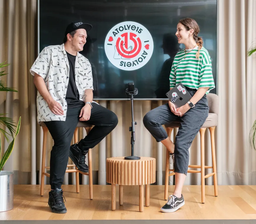 A man in a white shirt and a woman in a striped t-shirt smiling and sitting in front of a screen with the Atolye15 logo on it.