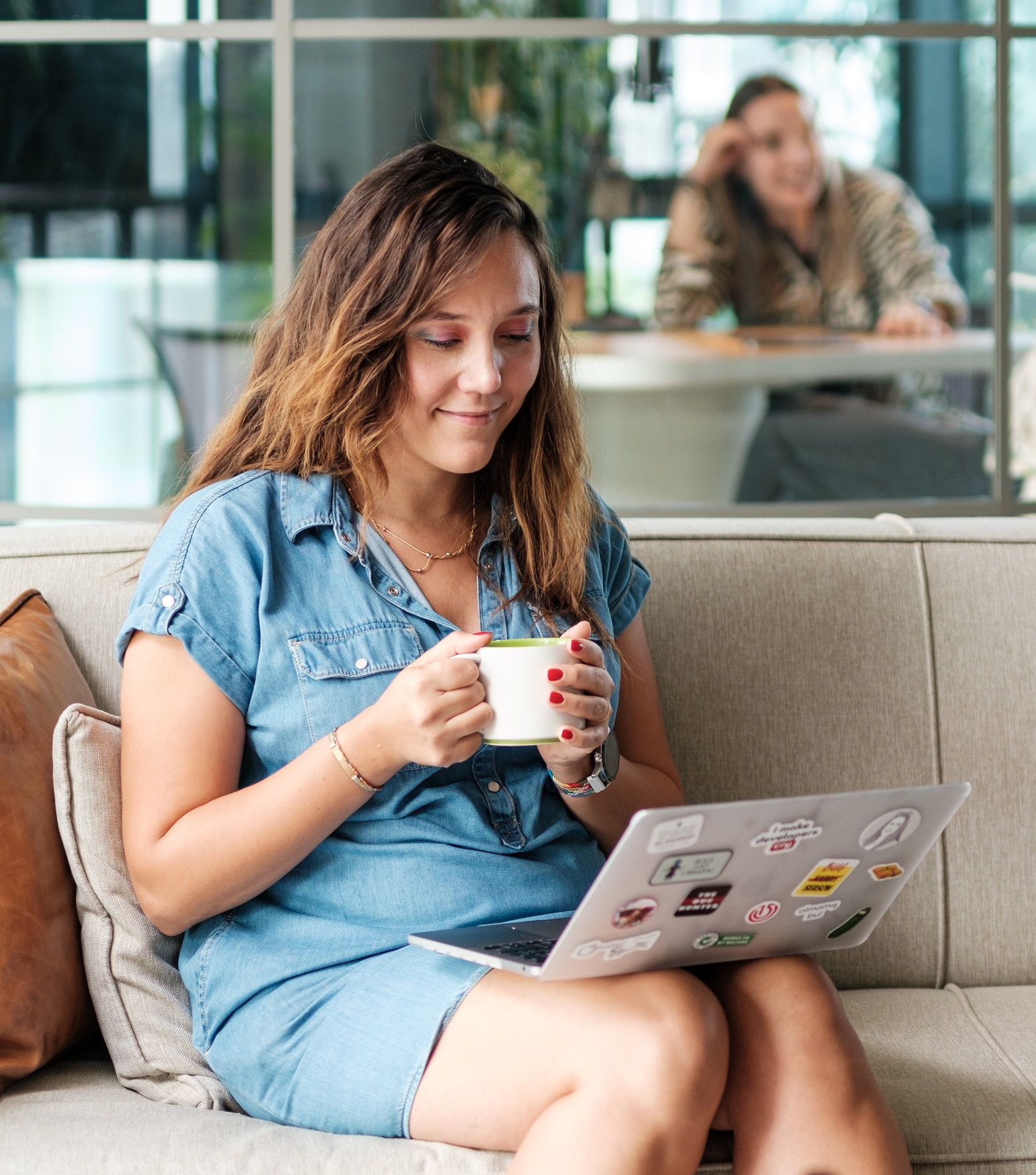 A smiling woman in a denim dress sitting on a couch with a white coffee mug and looking at a MacBook pro covered in stickers.